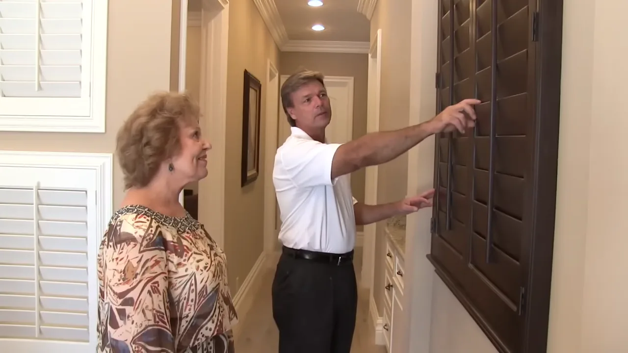 Golden West owner John showing a dark wood plantation shutter to a client in a home hallway, pointing out the louvers while she looks on