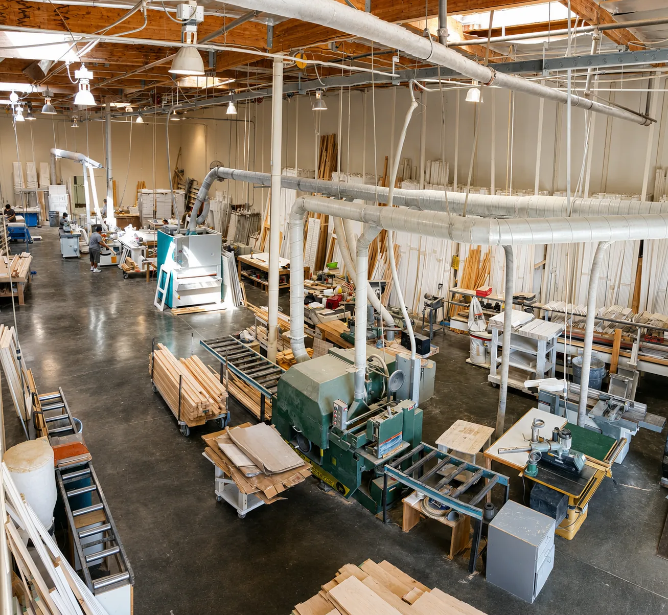 An elevated view of the Golden West Shutters factory floor in Lake Forest, with woodworking machinery, dust-collection ducting, stacked basswood and shutter components, and workers building custom shutters